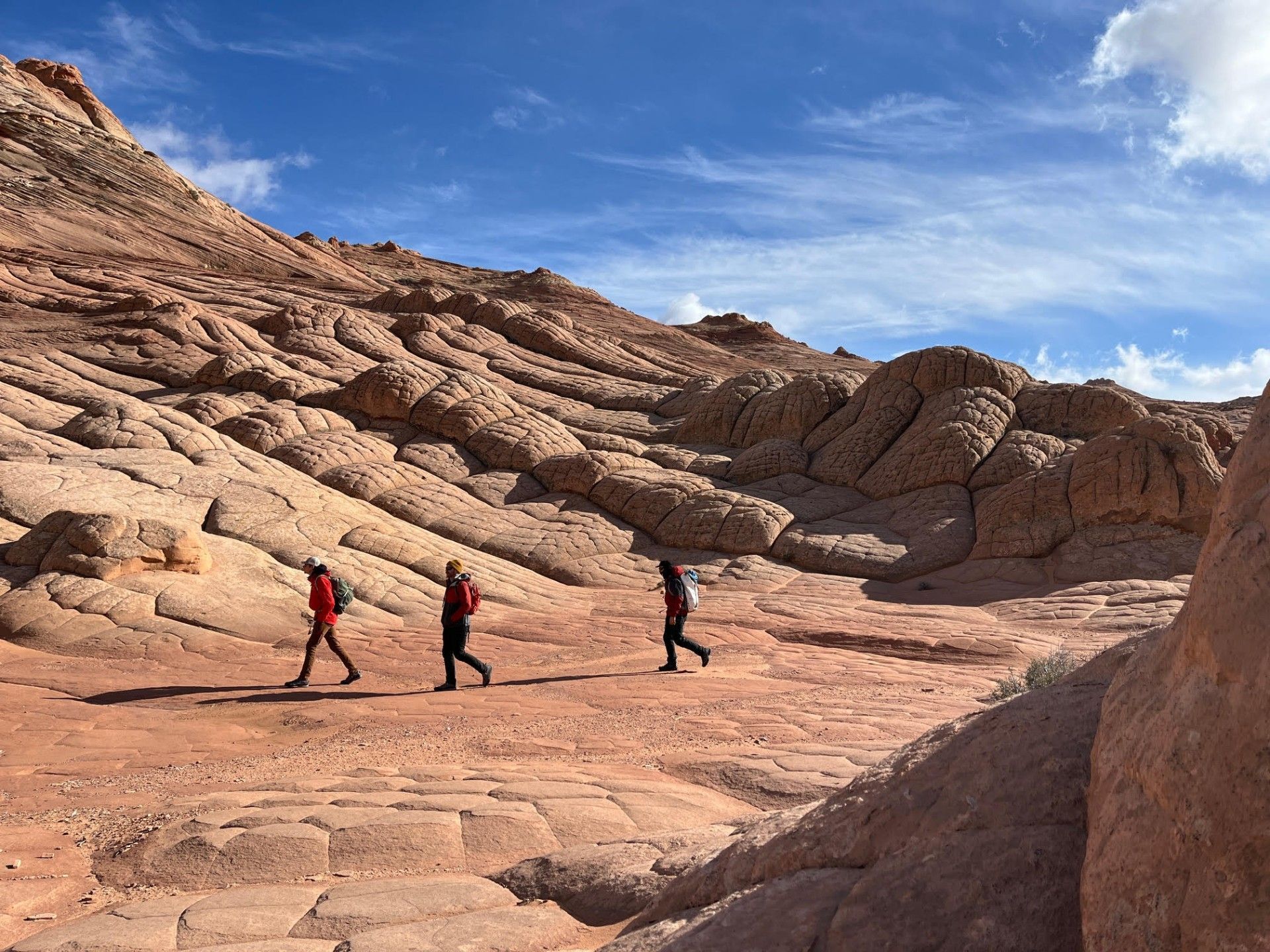 Three people hiking in red jackets through a red rock landscape under a blue sky.