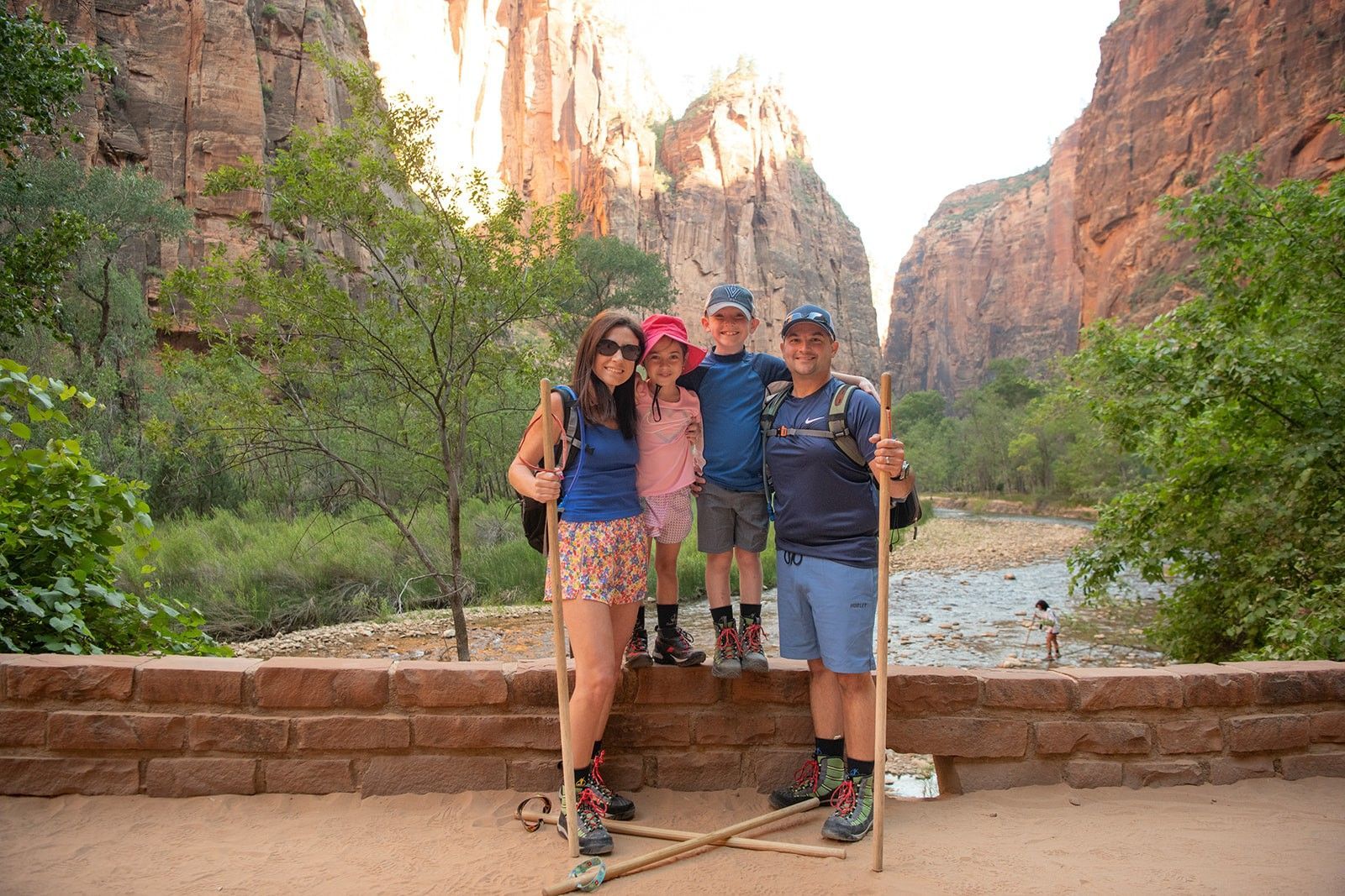 Family poses for photo near a river in a canyon. They hold walking sticks and smile.