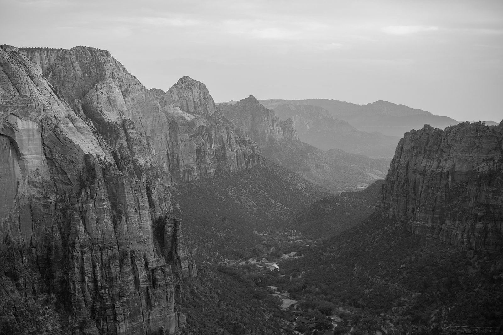 Black and white view of a canyon valley with rugged rock formations, trees, and a hint of buildings in the distance.