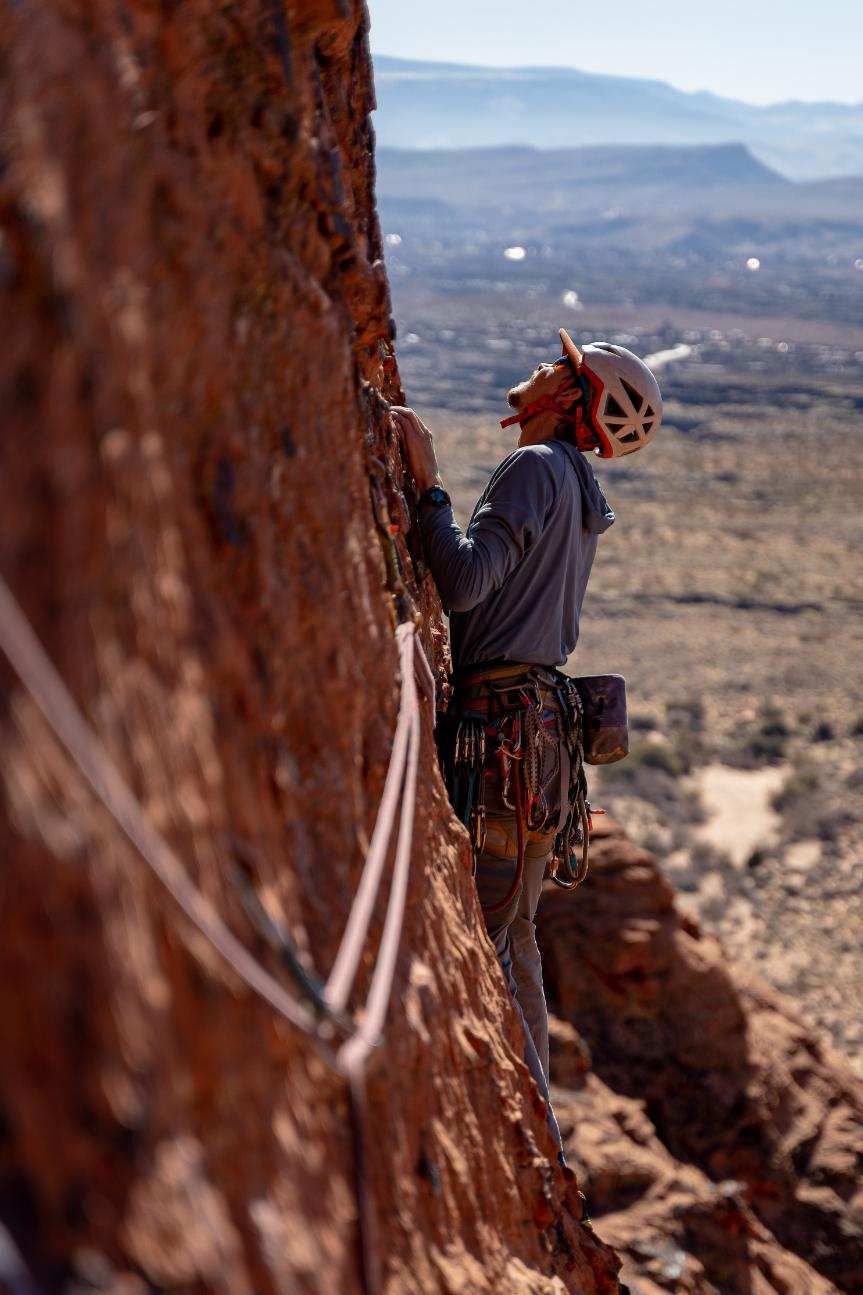 Rock climber ascending a red rock face, wearing a helmet and climbing gear, with a desert landscape in the background.