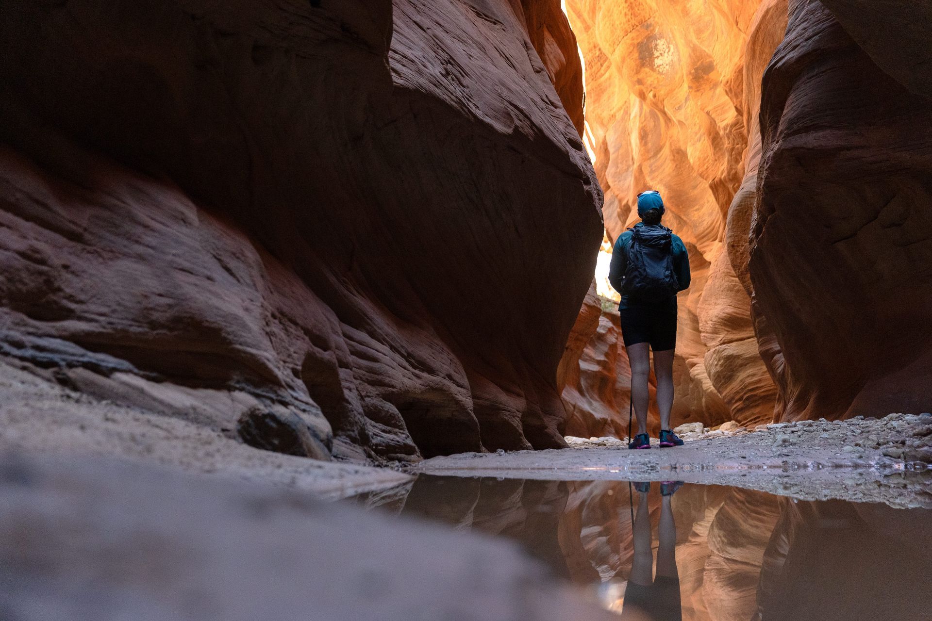 Person standing in a narrow slot canyon with reflections in a puddle; orange and brown rock walls.
