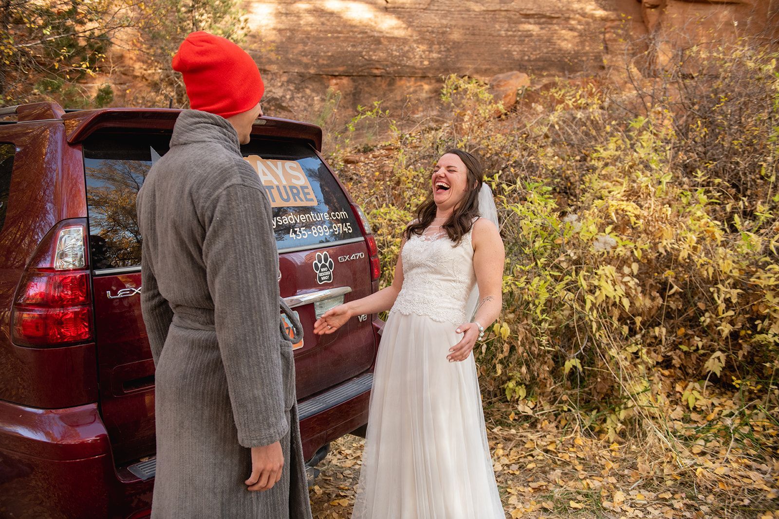 Bride in wedding dress laughs at person in robe and orange hat next to a car. Autumn setting.