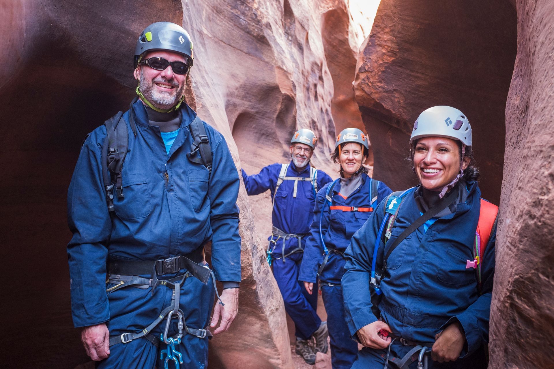 Four climbers in blue gear smiling in a narrow canyon, wearing helmets and harnesses