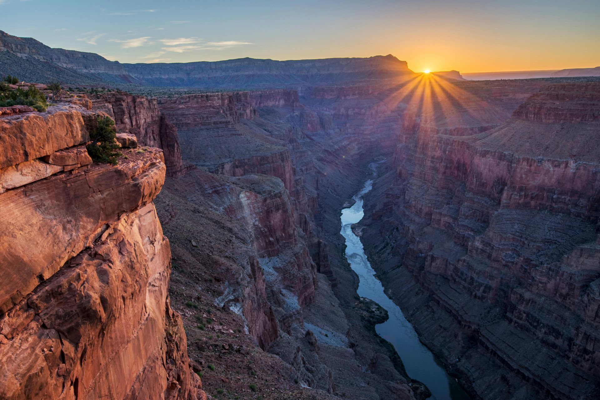 Sunrise over a deep canyon with a river, reddish-brown cliffs, and a bright sunburst.