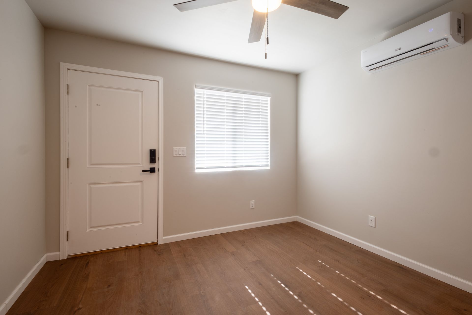 Empty beige bedroom with wood floor, white door, window blinds, ceiling fan, and wall-mounted AC unit