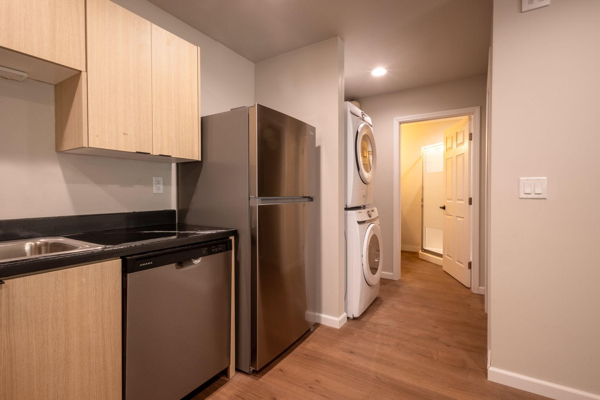 Compact kitchen with stainless steel appliances, light wood cabinets, and a hallway leading to a lit doorway