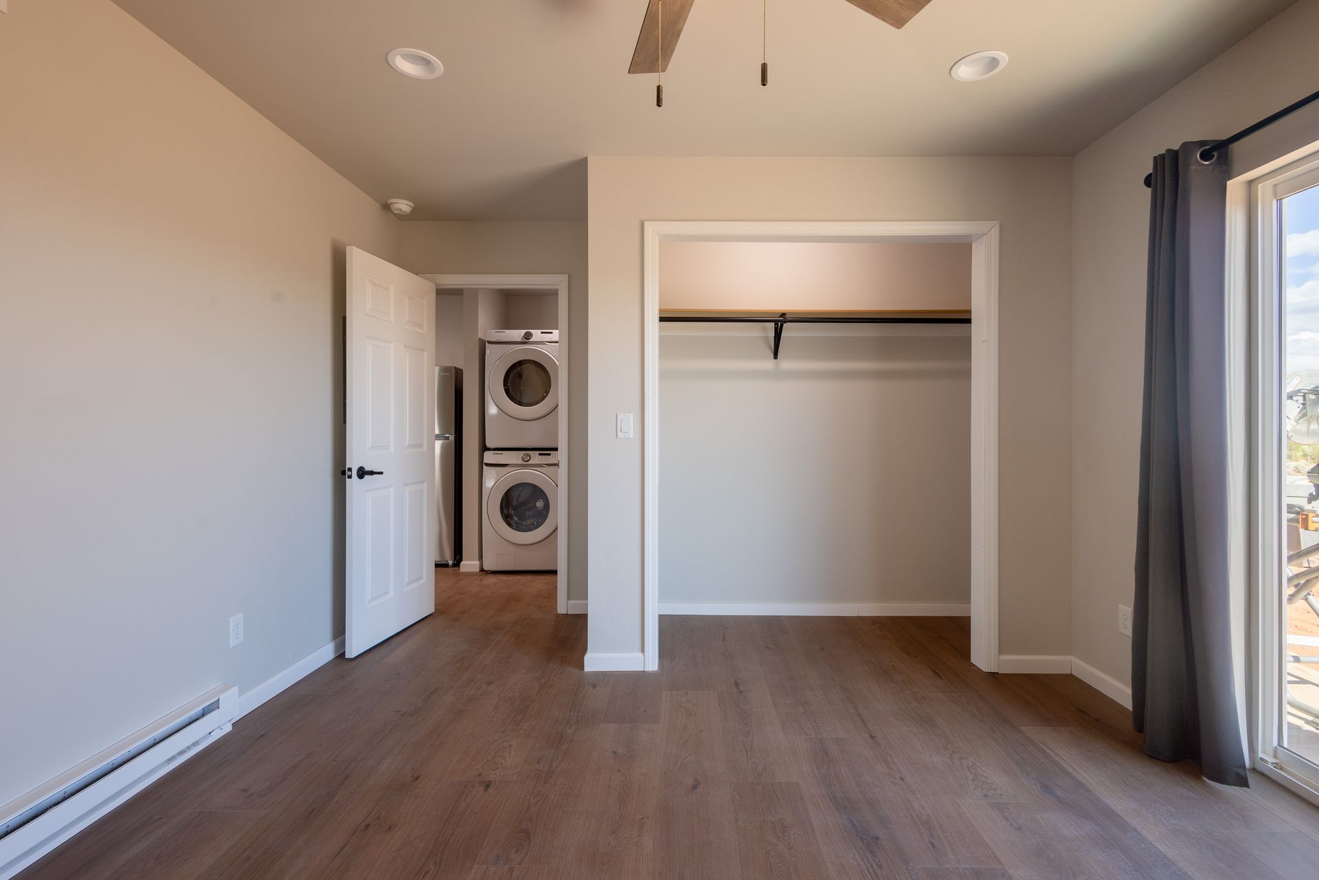 Empty beige bedroom with open closet, hardwood floor, and stacked washer-dryer in the hall.
