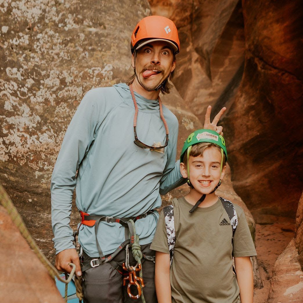 Man and boy in canyon, both wearing helmets, man making a playful face, boy smiling.