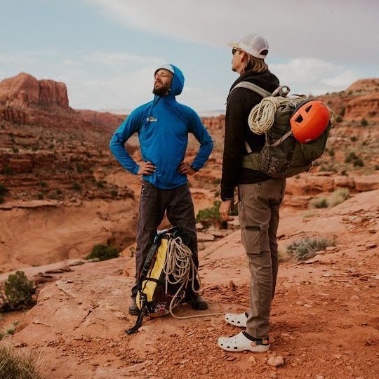 Two hikers in a desert landscape. One in blue, looking up; the other with a backpack and orange helmet, looking at him.