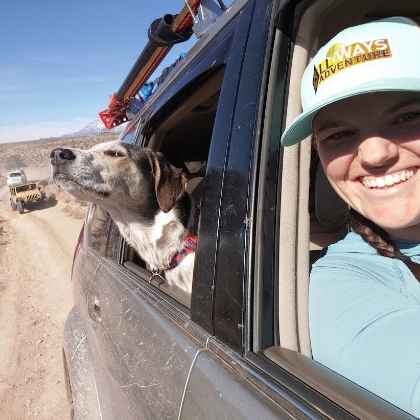 A woman smiles, taking a selfie with her dog in a car on a dusty road; mountains and a vehicle are in the background.