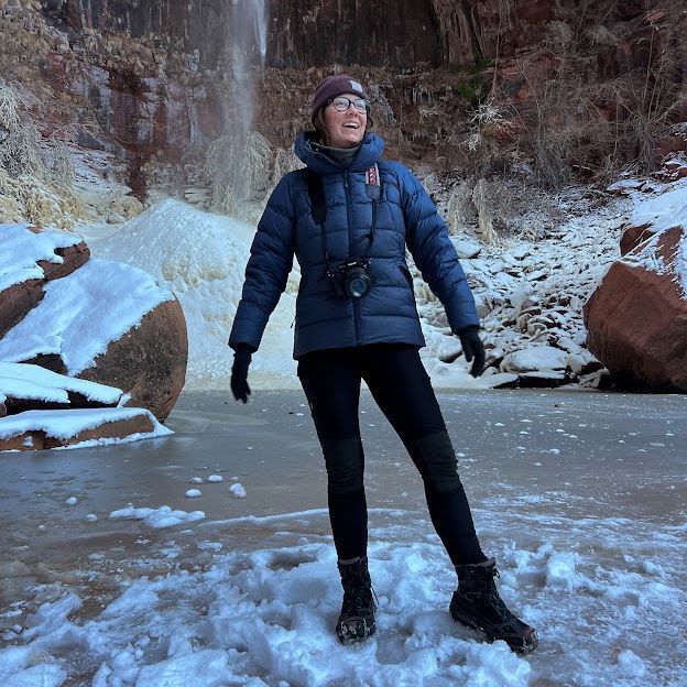 Woman in winter wear smiles in front of a waterfall and ice, with snow-covered ground and red rock.
