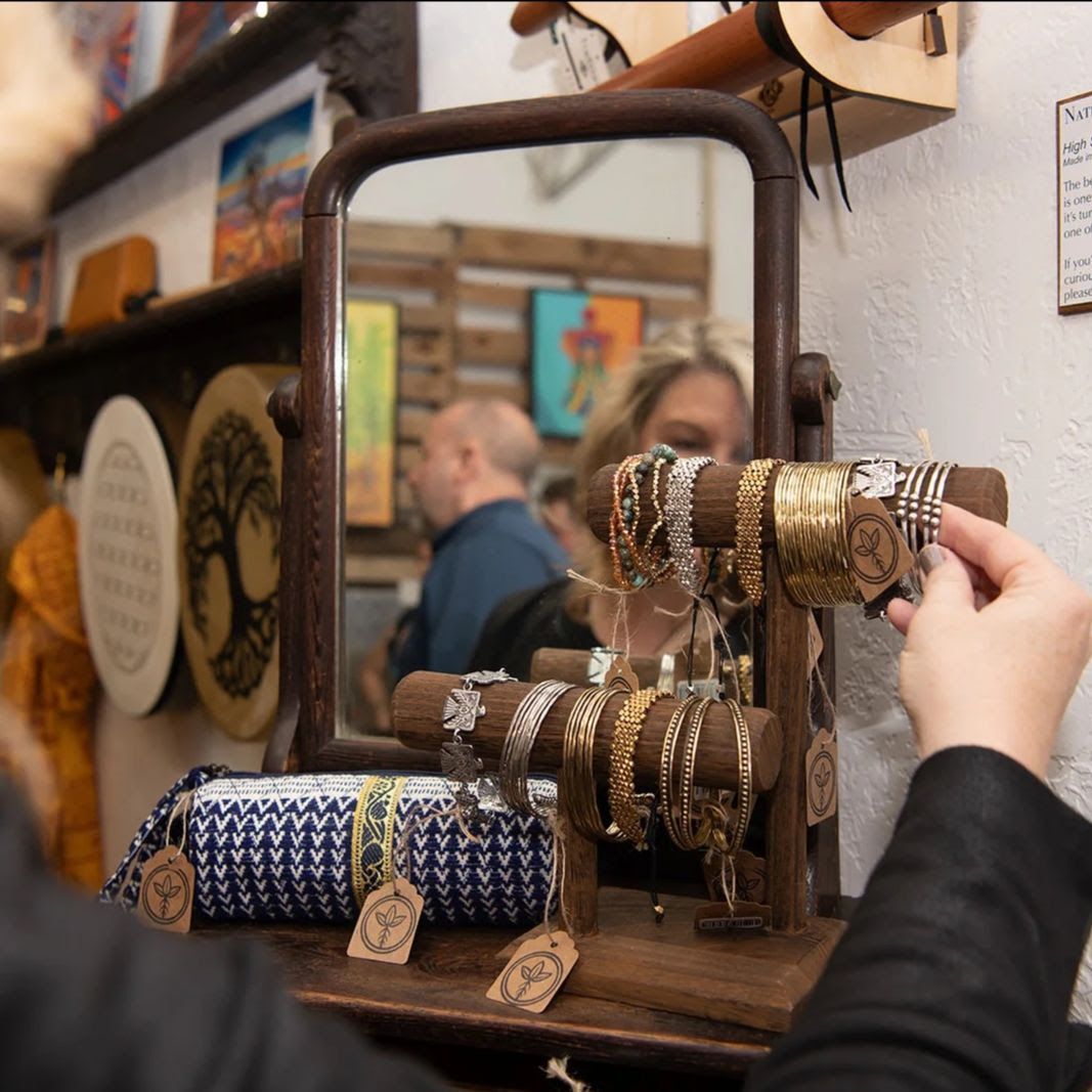 Person browsing jewelry on display in a shop. Mirror reflects other shoppers and art on shelves.