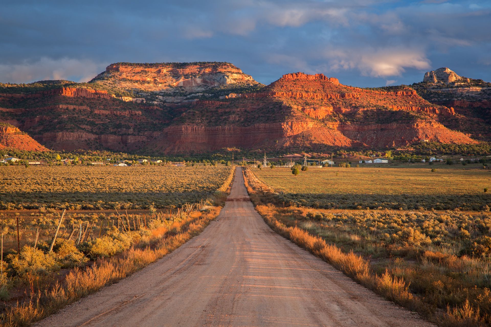 Dirt road leading towards reddish mountains at sunset. Fields flank the road under a blue sky.
