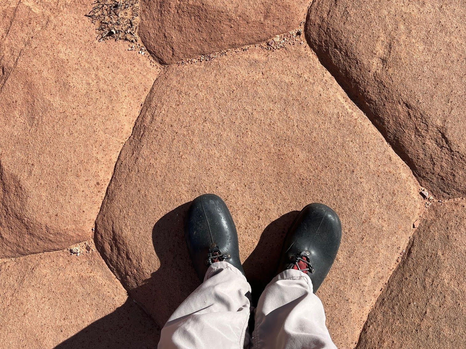 Feet in black shoes on reddish, tiled ground. Light-colored pants.