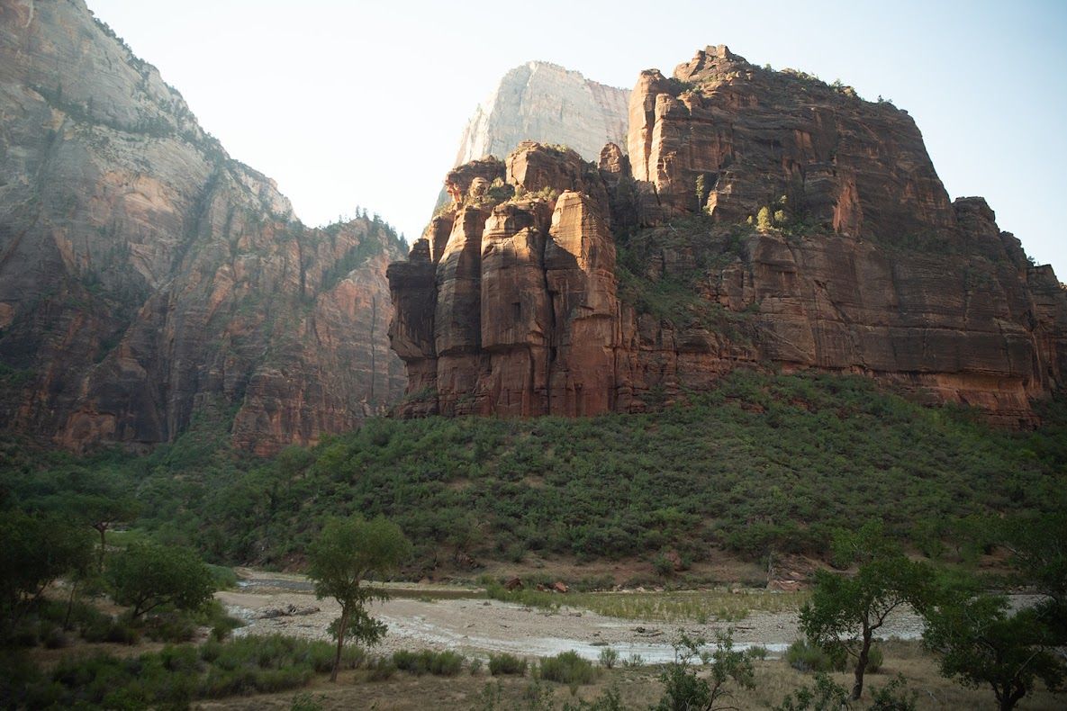 Red rock cliffs rise above green vegetation; a riverbed is in the foreground.