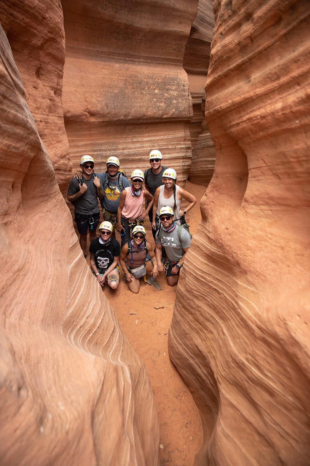 People in raft on river, smiling. Red-rock canyon walls rise on either side. Cloudy sky.