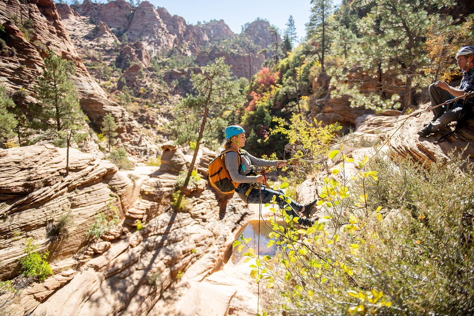 Woman rappels down a sandstone canyon, wearing a helmet and backpack, surrounded by trees and rocks.
