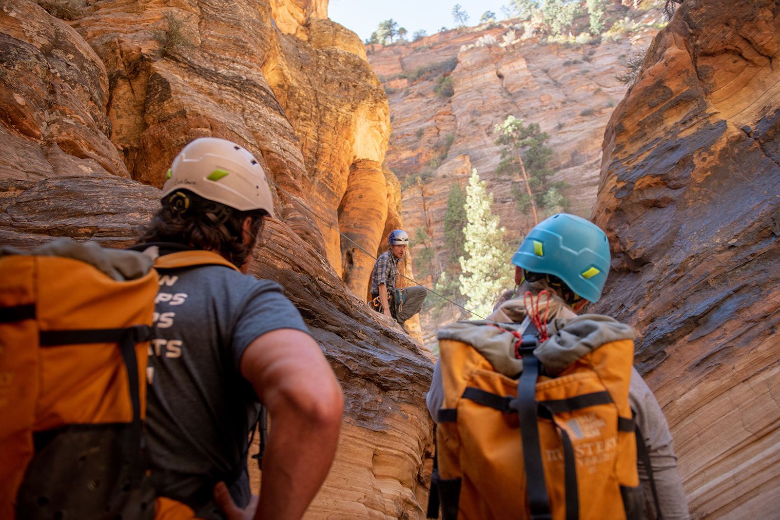 Hikers in canyon, wearing helmets and backpacks, looking up at others; rocky, sunlit environment.
