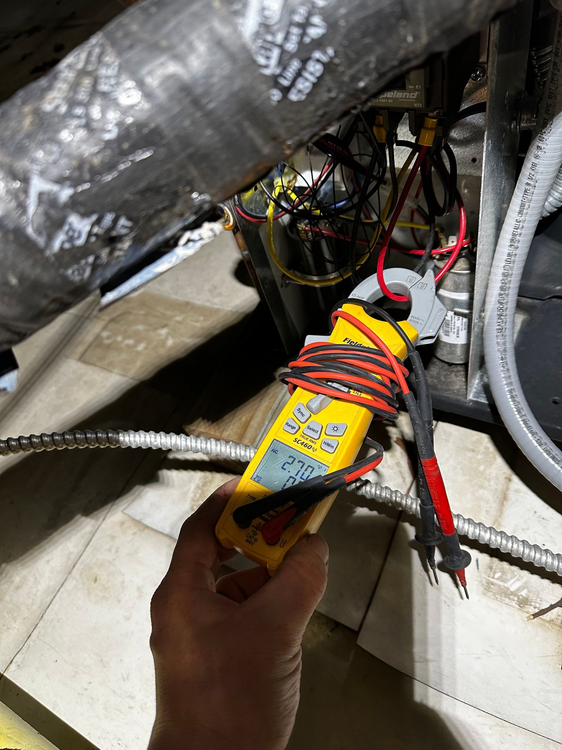 Hand holding a yellow multimeter with red and black probes testing electrical components near ductwork.
