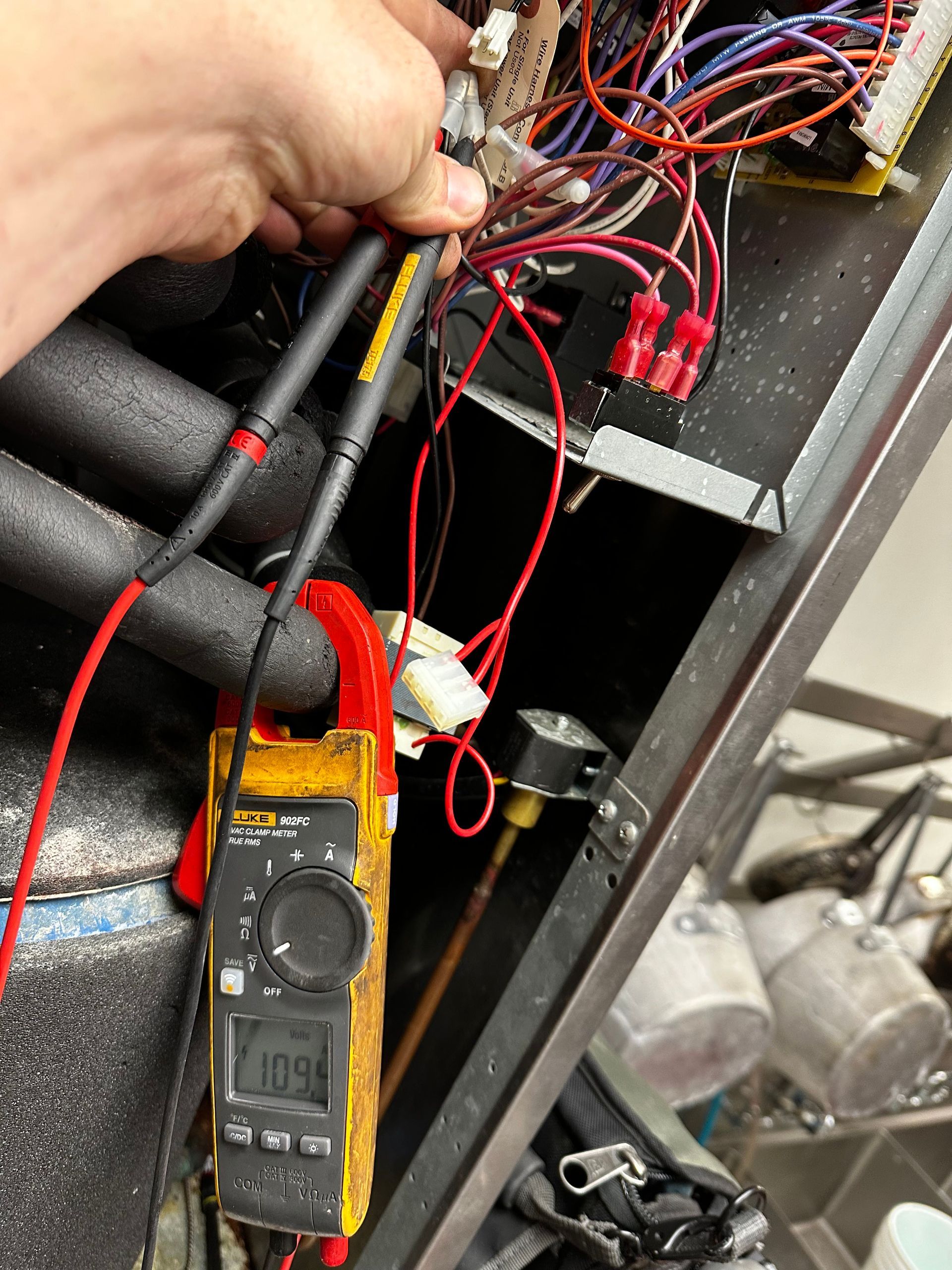 Hand using a multimeter to test electrical components inside an HVAC unit, showing a reading of 109.