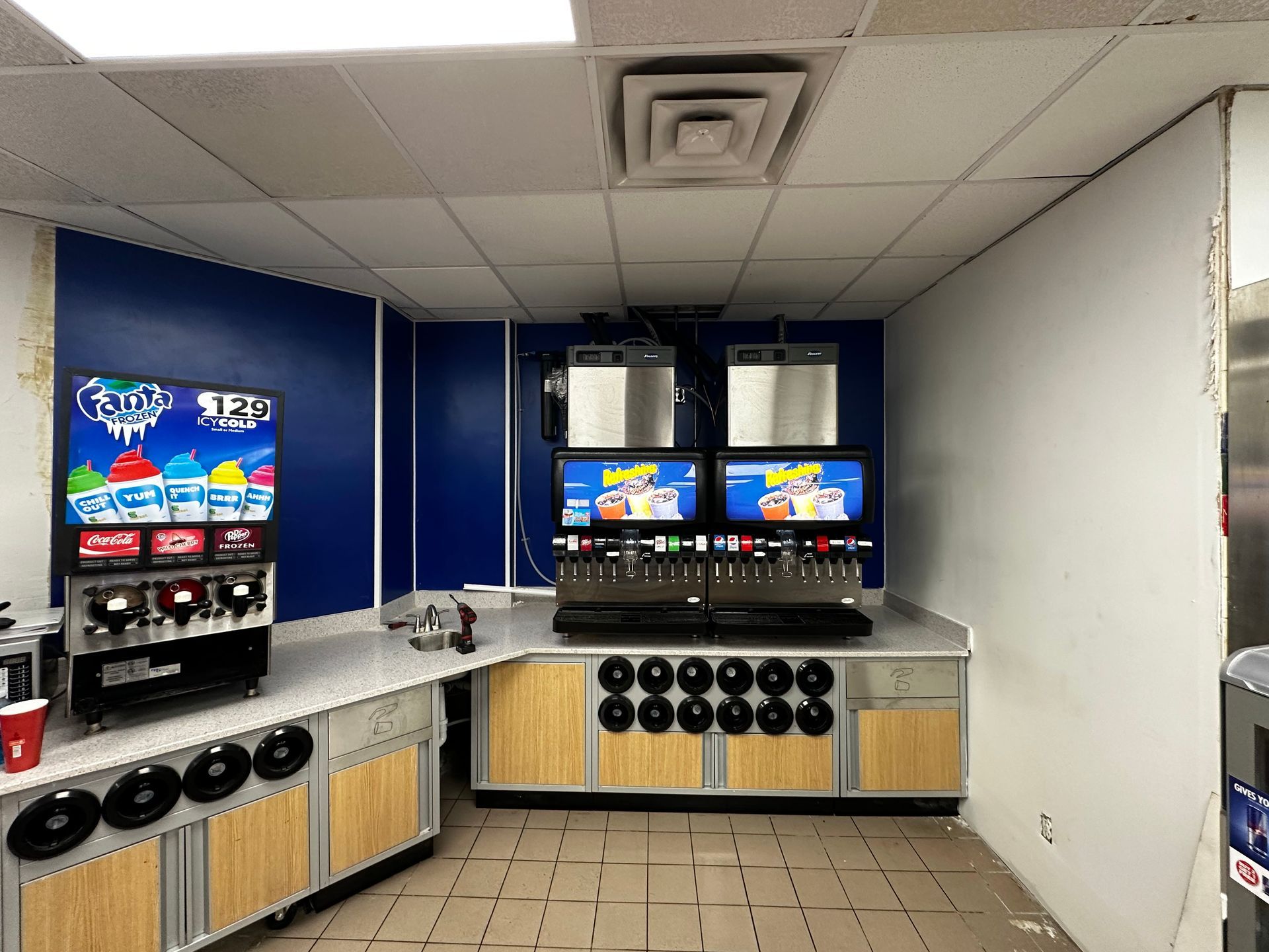 Soda fountain station with drink dispensers, blue and white walls, light tan cabinets.