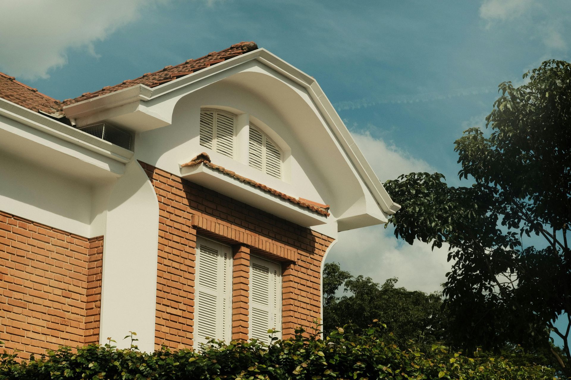 Brick house with white trim and arched roofline under a blue sky, seen above green trees