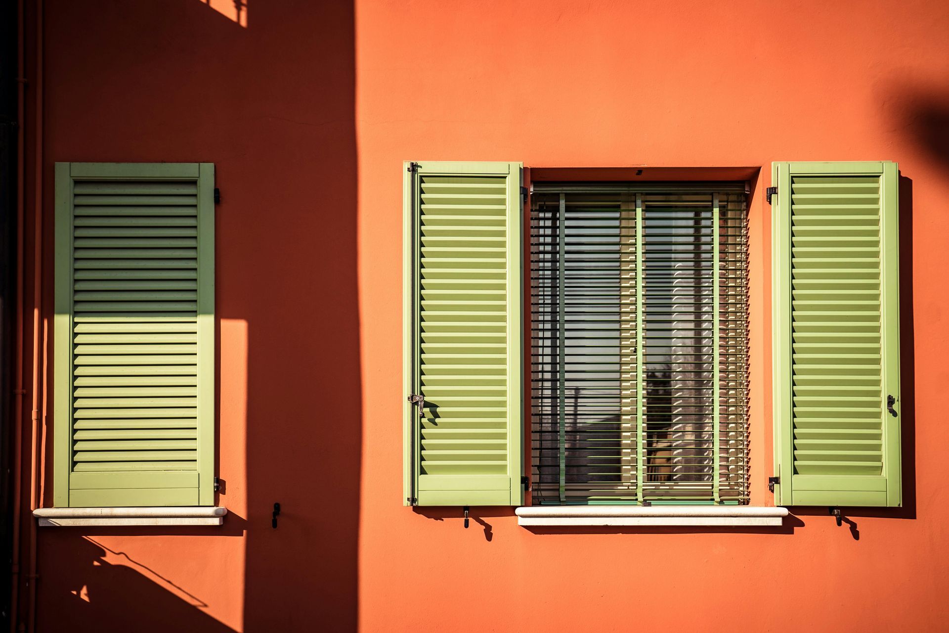 Orange wall with two green shuttered windows, one open and one closed, in bright sunlight