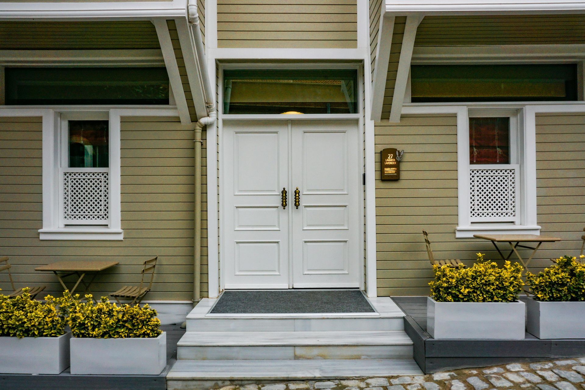 Front entrance of a beige house with white double doors, windows, steps, and flower boxes