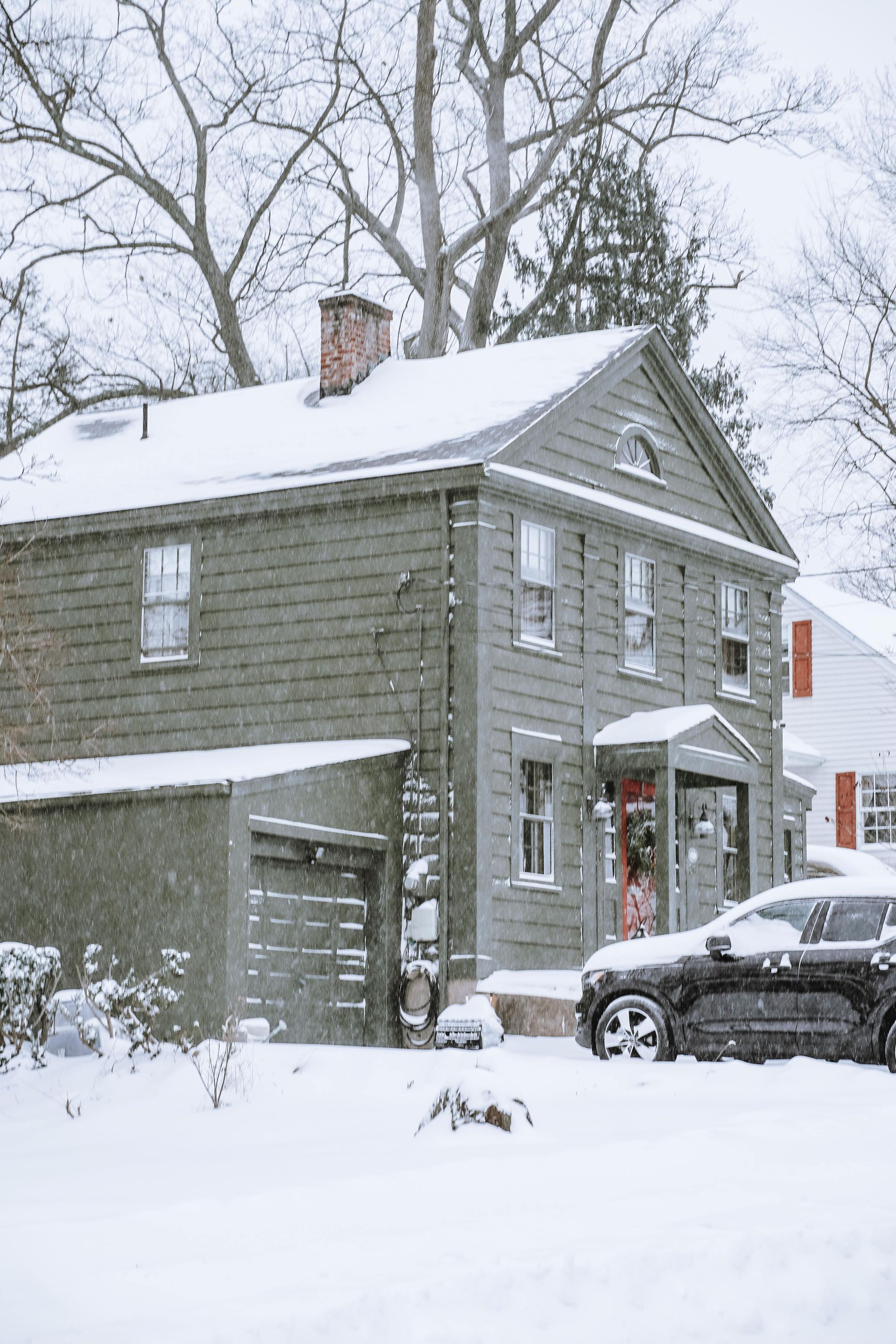 Snow-covered gray house with a parked car and bare trees during a winter storm