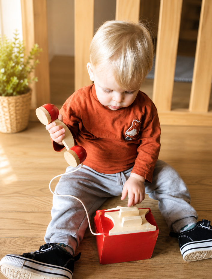 Toddler sitting on the floor, playing with a red toy phone in a wooden room.