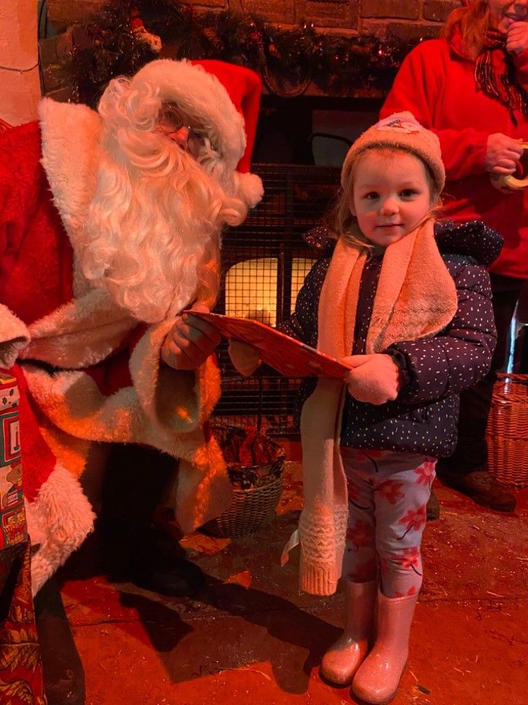 Child posing with Santa in a warmly lit holiday setting, wearing a scarf and patterned leggings.