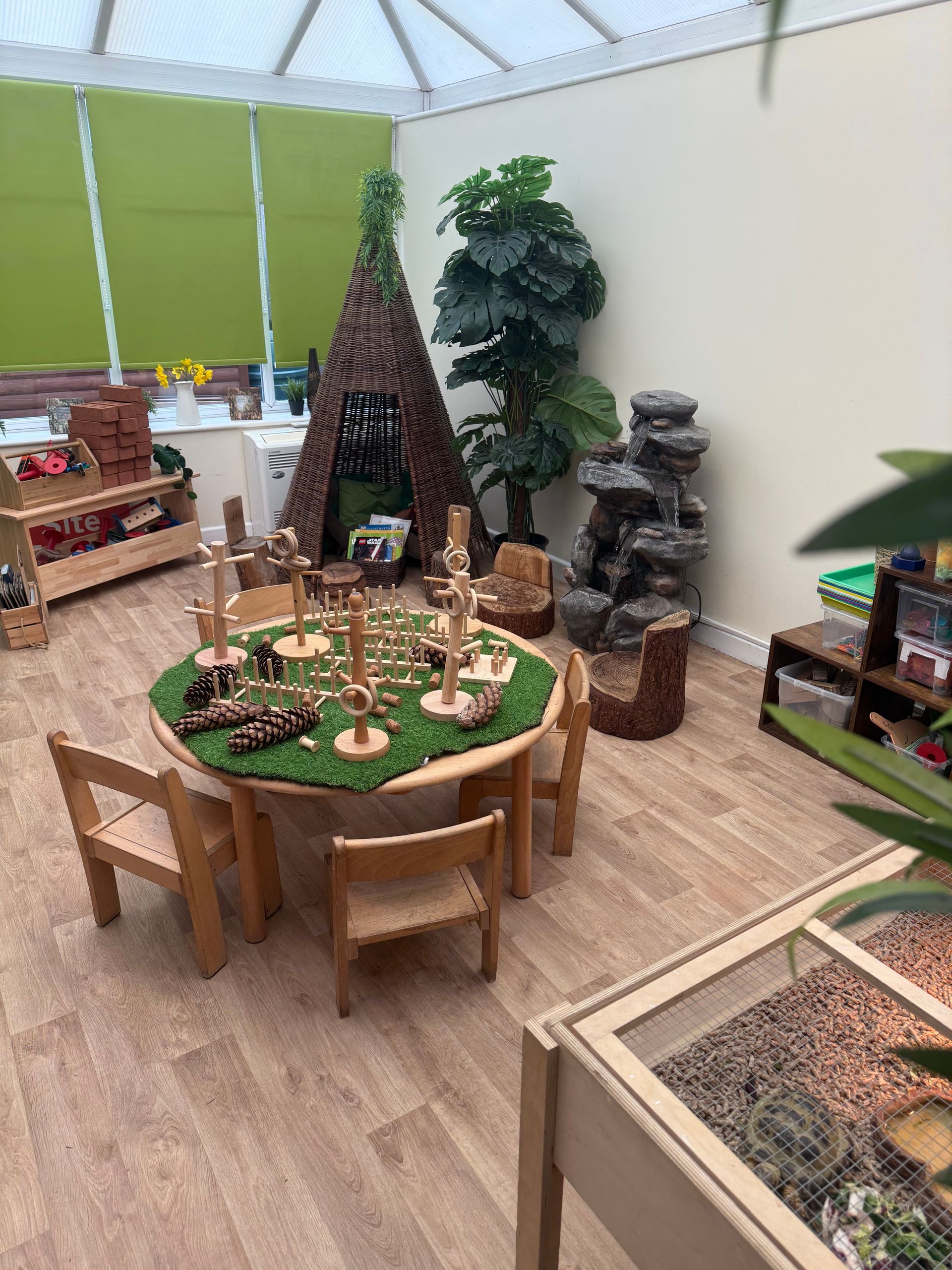 Indoor playroom with a round wooden table, chairs, toy garden display, and small play structures.