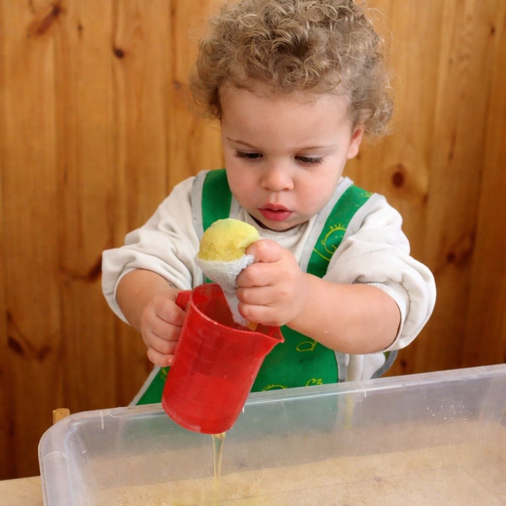 Child in a green apron pouring from a red cup into a clear bin, holding a yellow scoop.