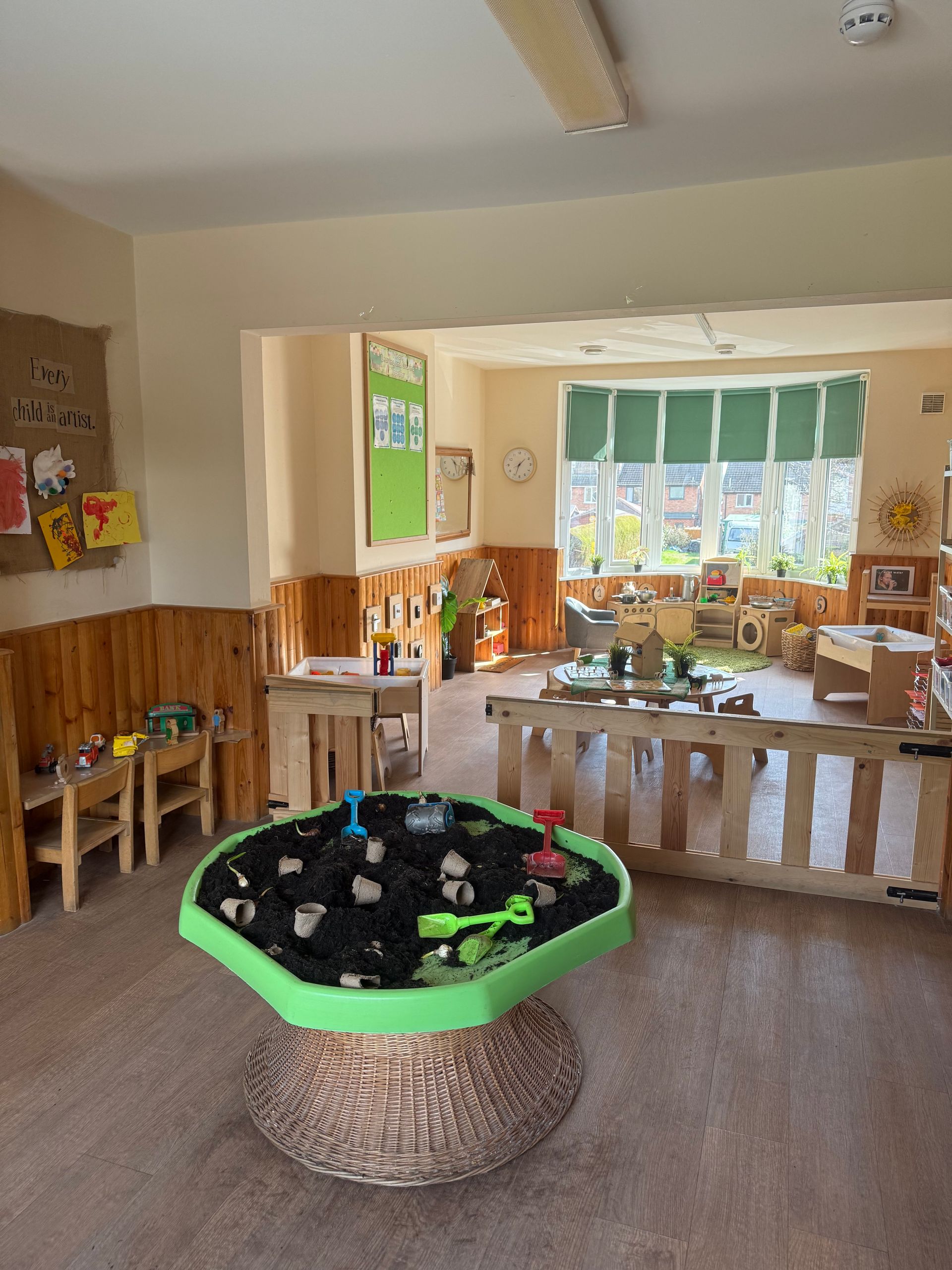 Bright preschool classroom with wooden furniture, a round activity table, and colorful window decor.