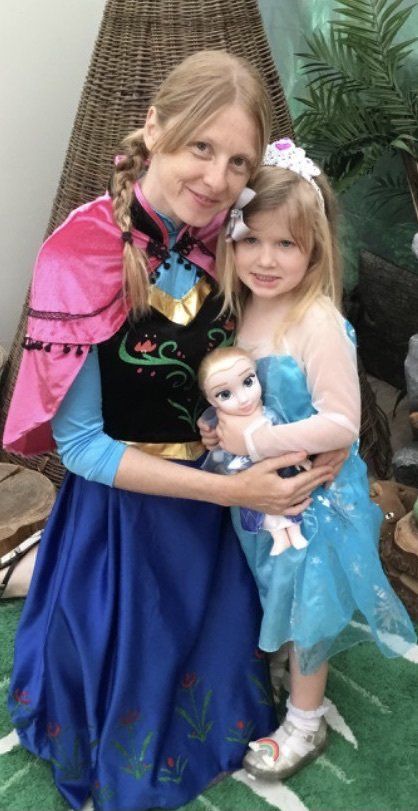 Woman and young child in costumes holding a doll, posing outdoors with greenery behind them