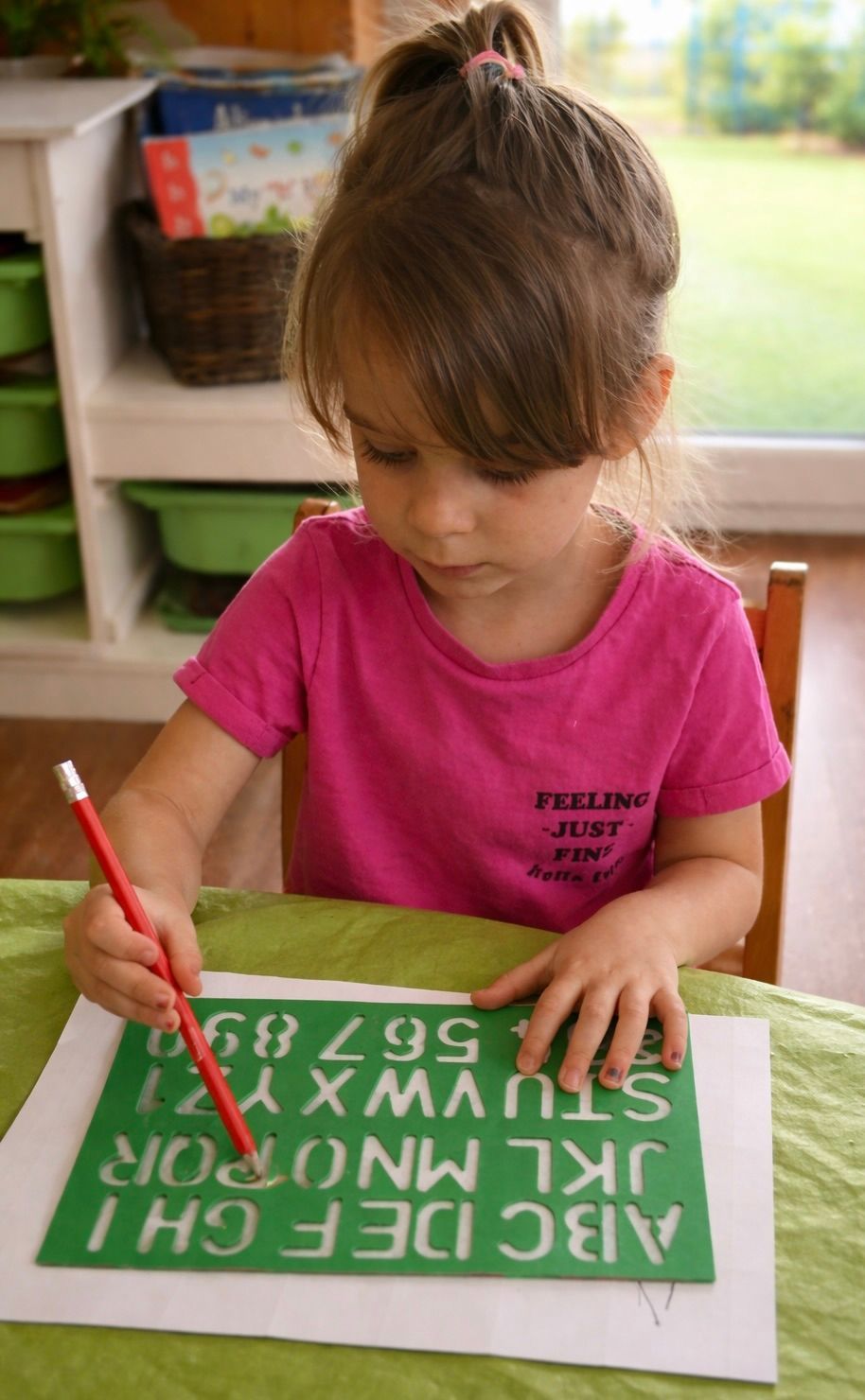Child in pink shirt tracing letters on a green alphabet board with a red pencil