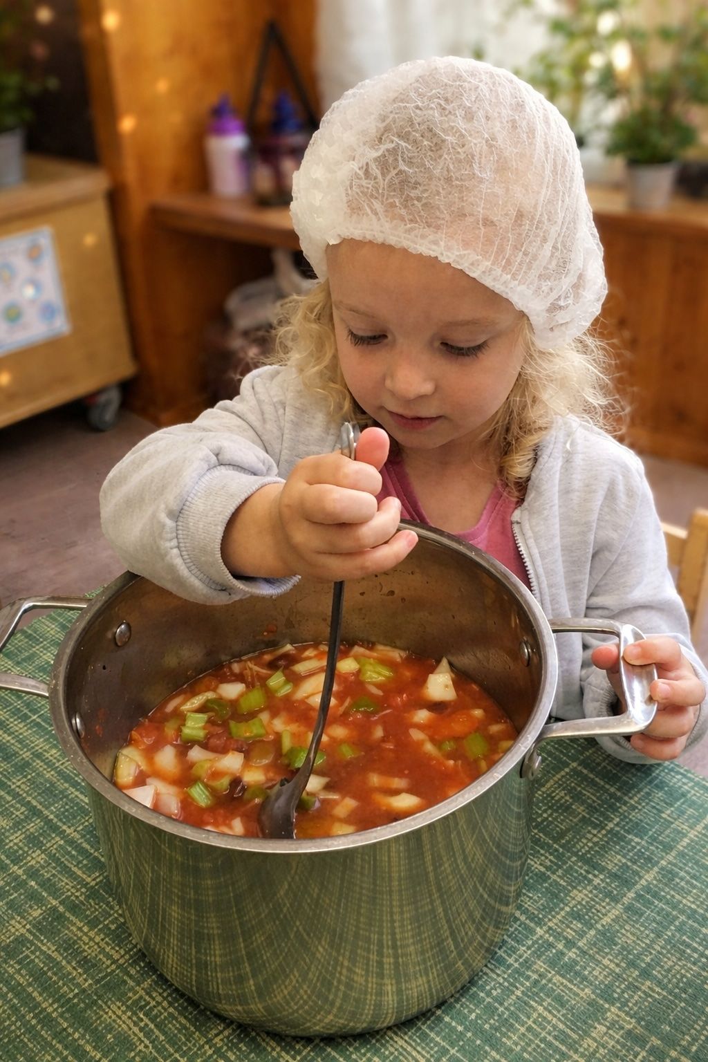 Young child stirring a pot of colorful soup in a kitchen, wearing a hair net and white sweater