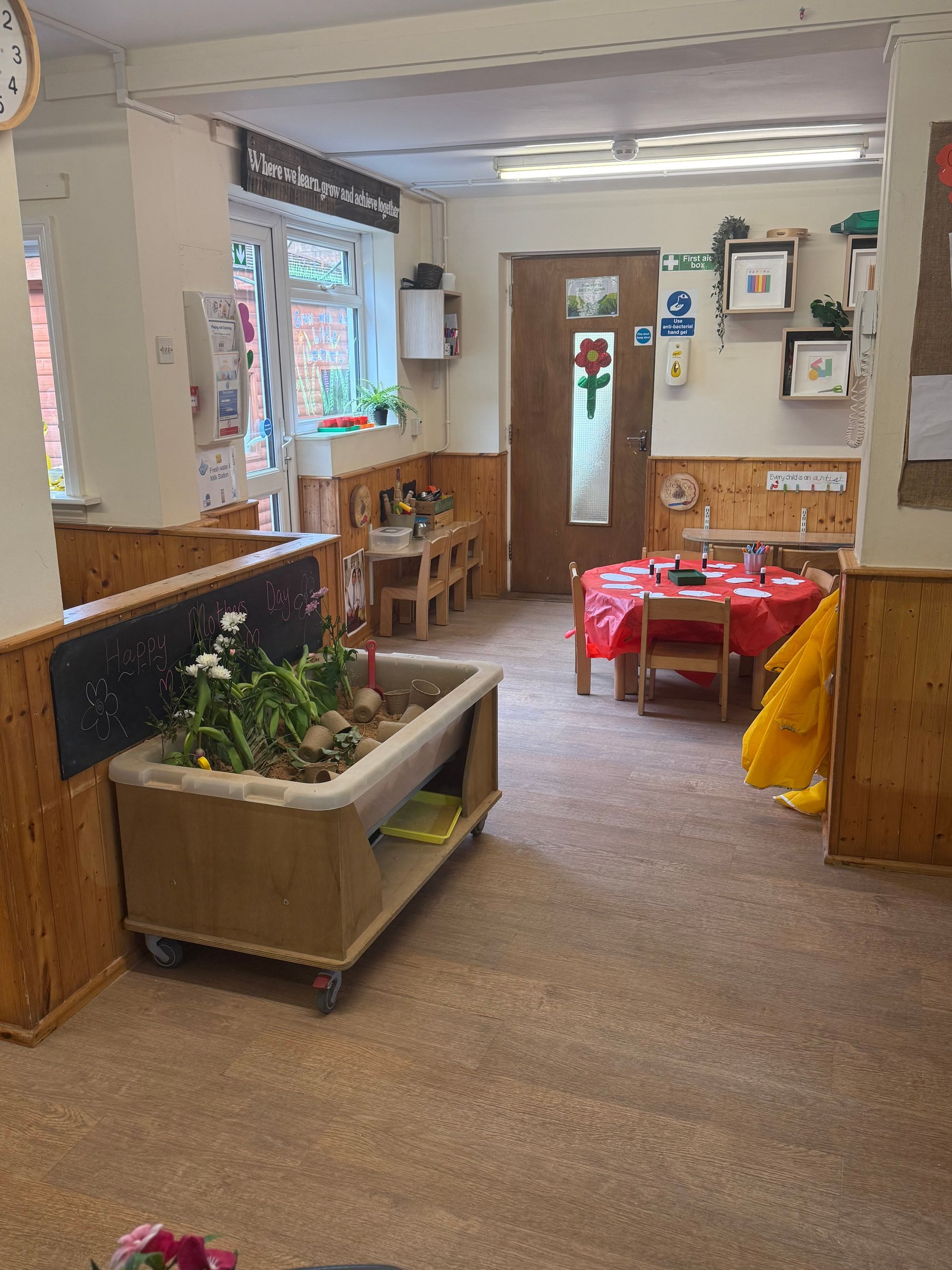 Bright daycare room with tables, chairs, toys, and a small play area near windows and cabinets