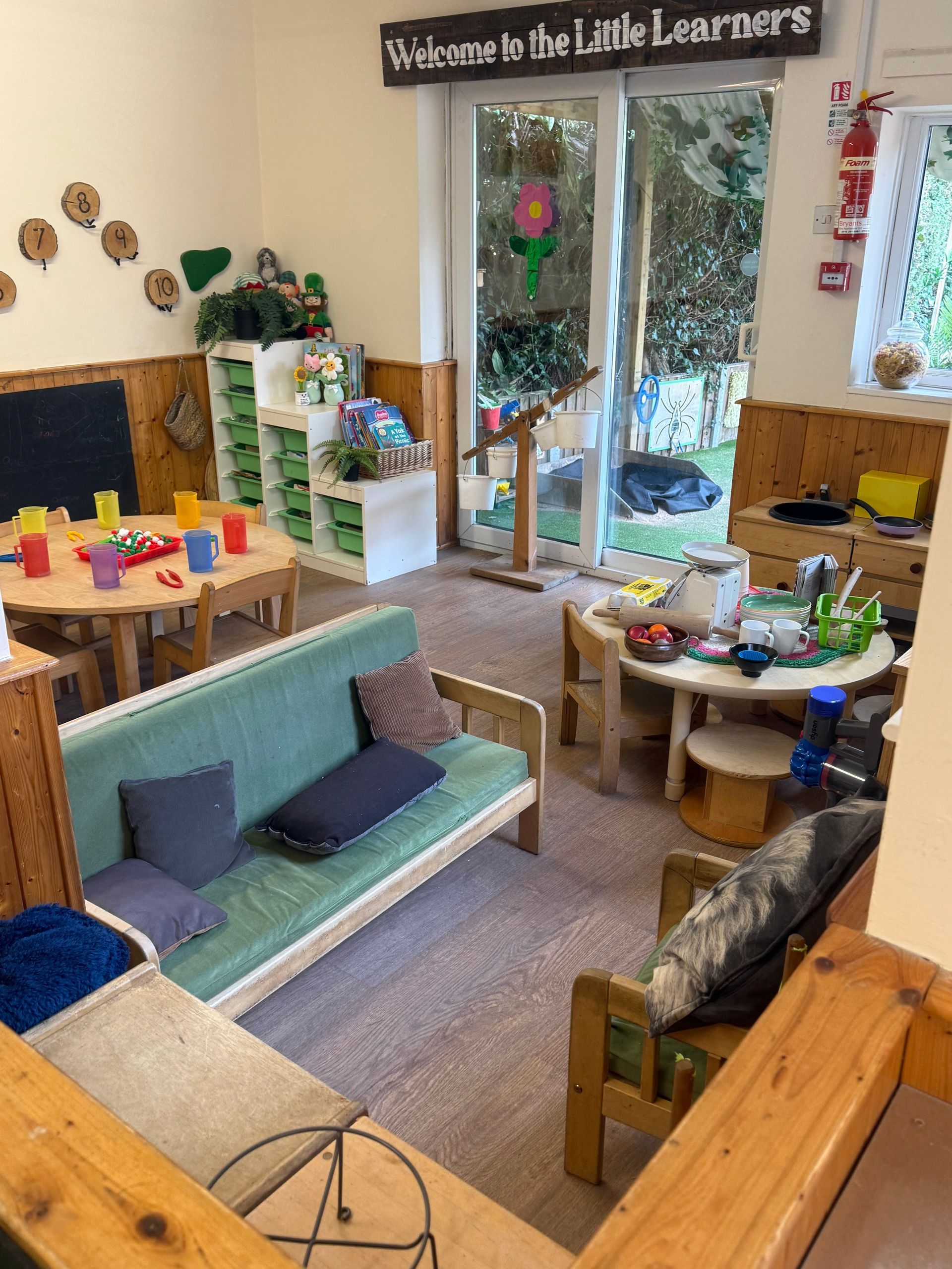 Bright children’s playroom with toy shelves, small tables, and a sofa by glass doors to a garden