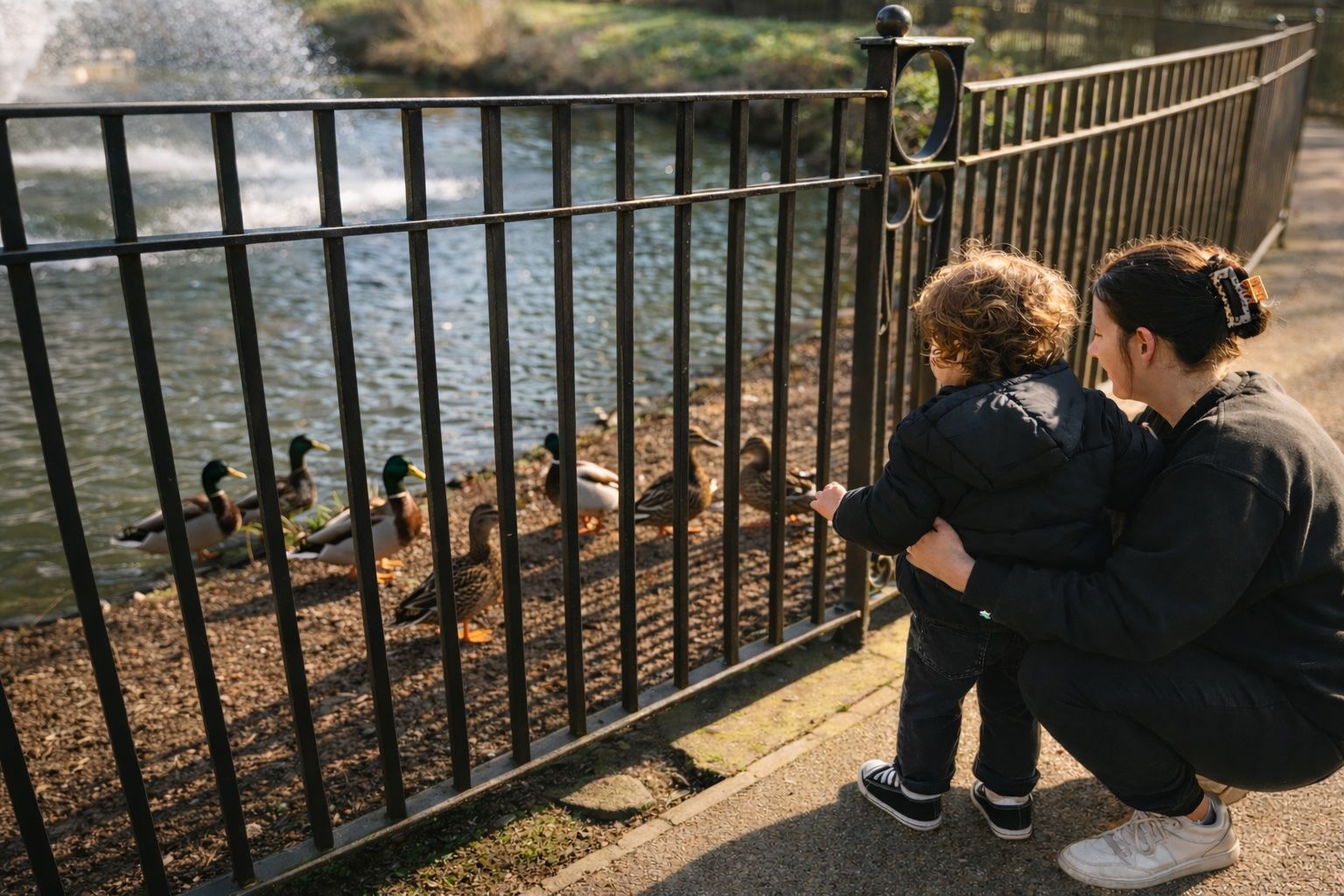 Adult and child crouch by a black fence, watching ducks in a sunlit park pond.
