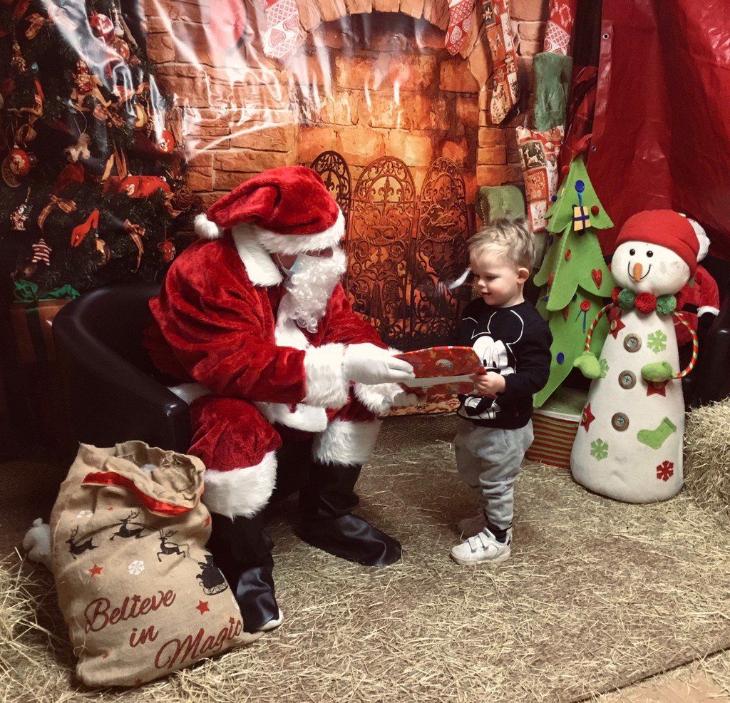 Santa handing a gift to a toddler in a festive holiday display with red decorations and a snowman figure