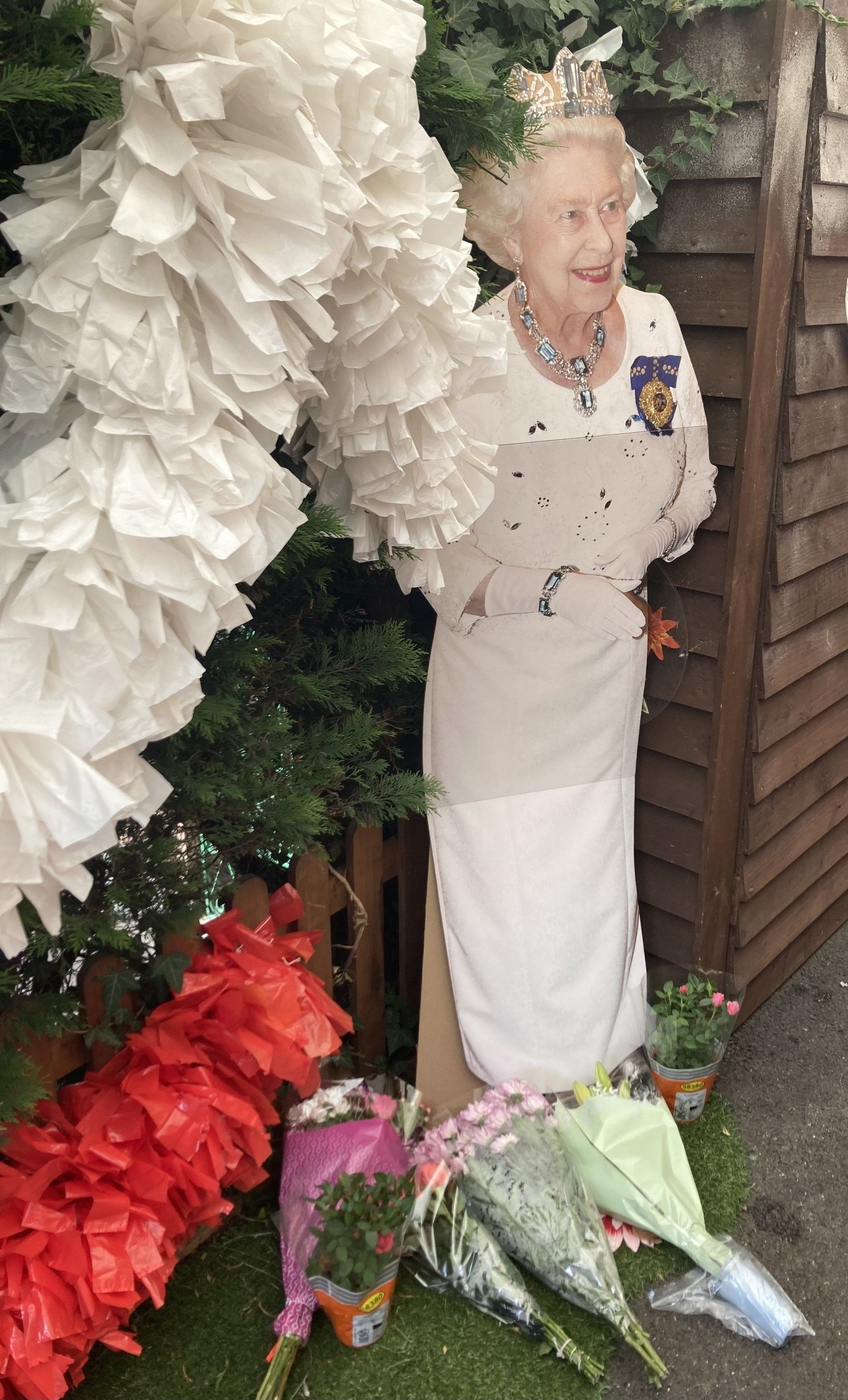 Cardboard cutout of Queen Elizabeth II beside white and red floral displays and bouquets outdoors