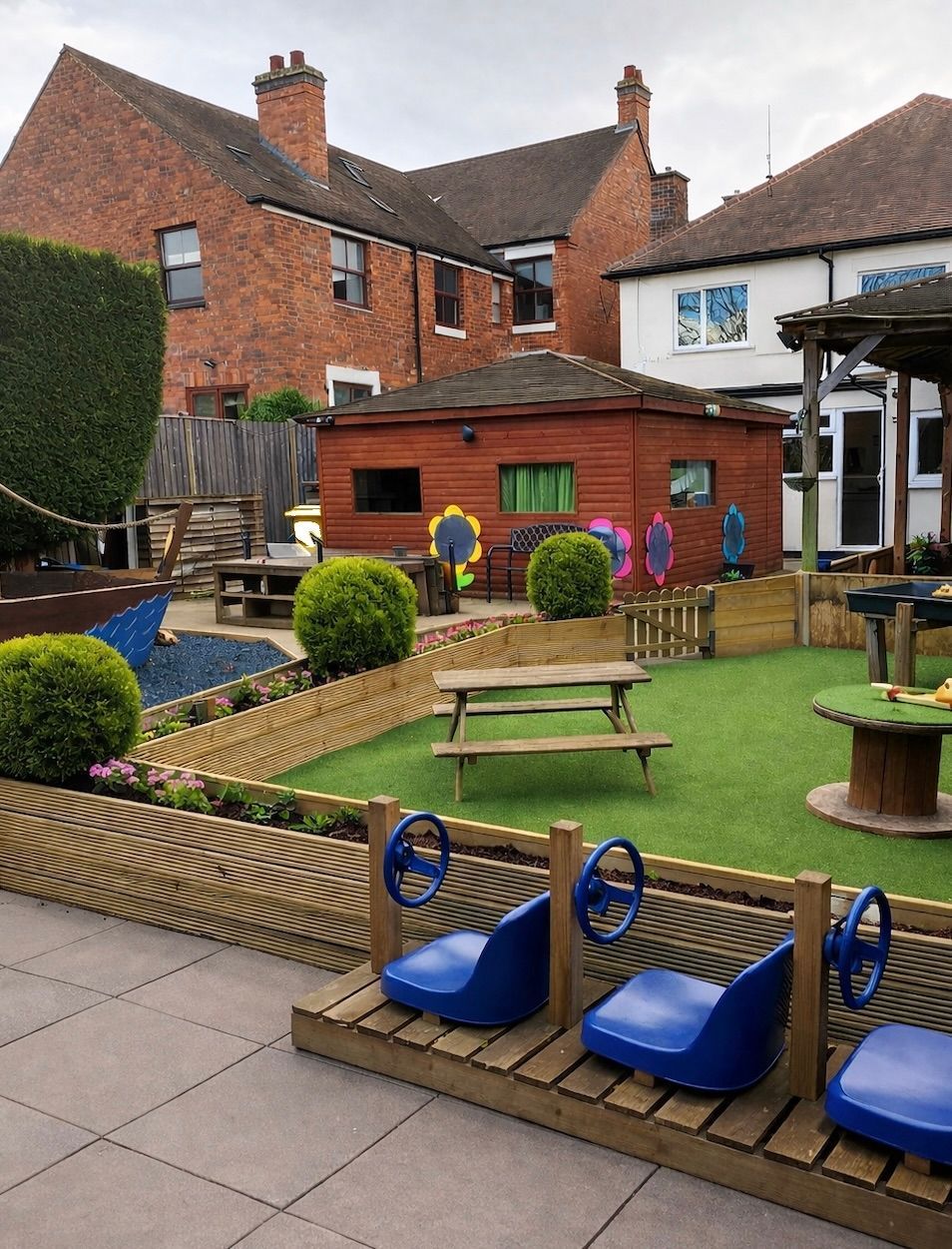 Small playground with blue spring riders, benches, and a grassy yard beside brick buildings.