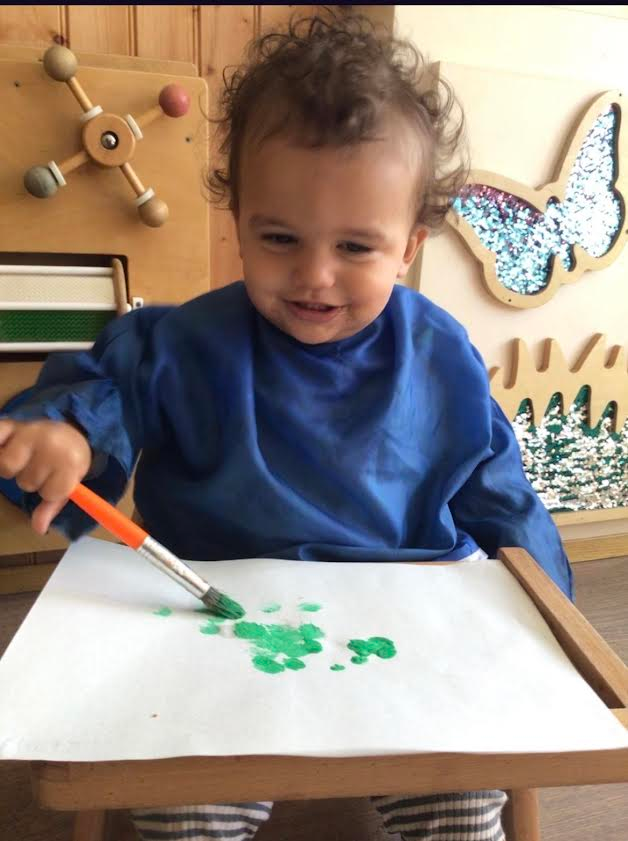 Child painting green dots on paper at a table, wearing a blue smock.