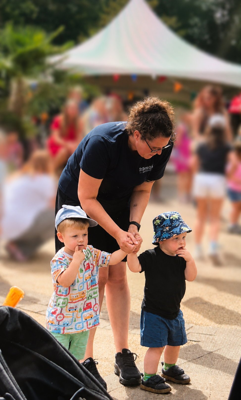 Adult helping two toddlers walk on a sandy beach, with a white tent and blurred crowd in the background.