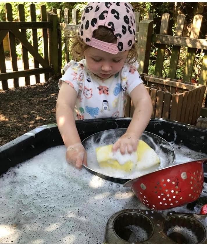 Child washing dishes in a tub outdoors, wearing a pink leopard-print hat.