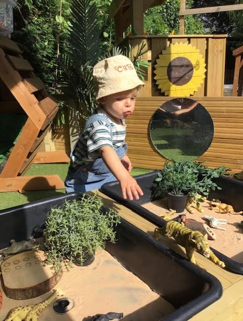 Child in a sun hat playing with sand and plants in a wooden outdoor sensory garden area.