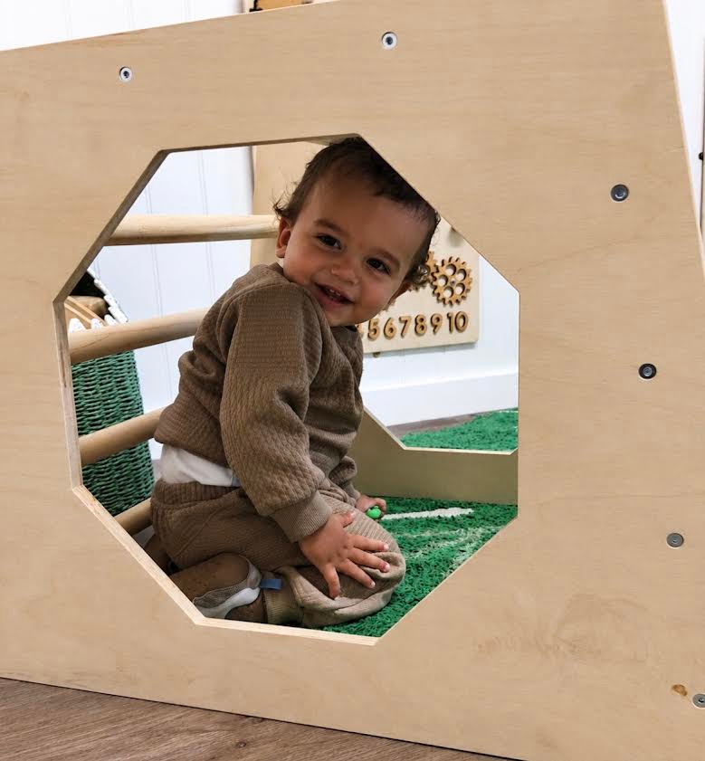 Smiling toddler peeks through an octagonal cutout in a wooden play panel.