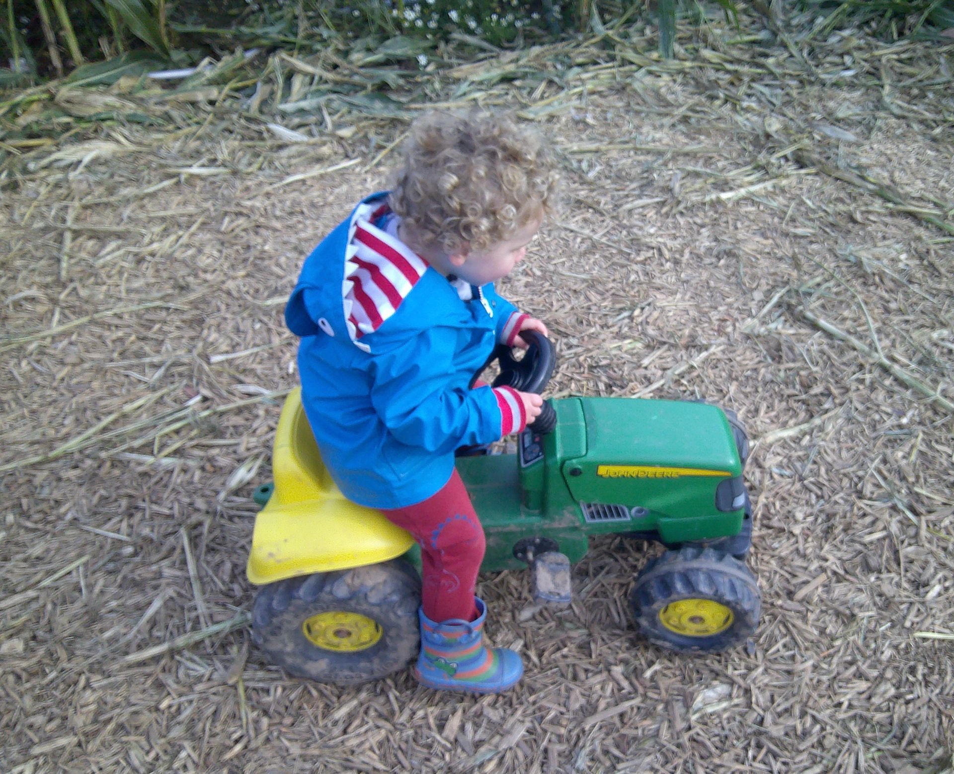 Child riding a small green toy tractor outdoors on a dirt-and-straw ground