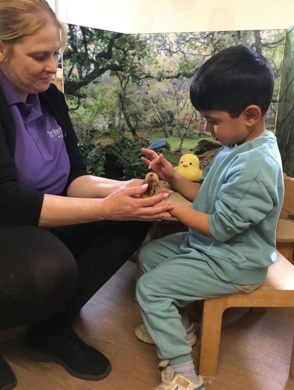 Adult and child sharing a small bird in a nature setting, seated beside a forest backdrop